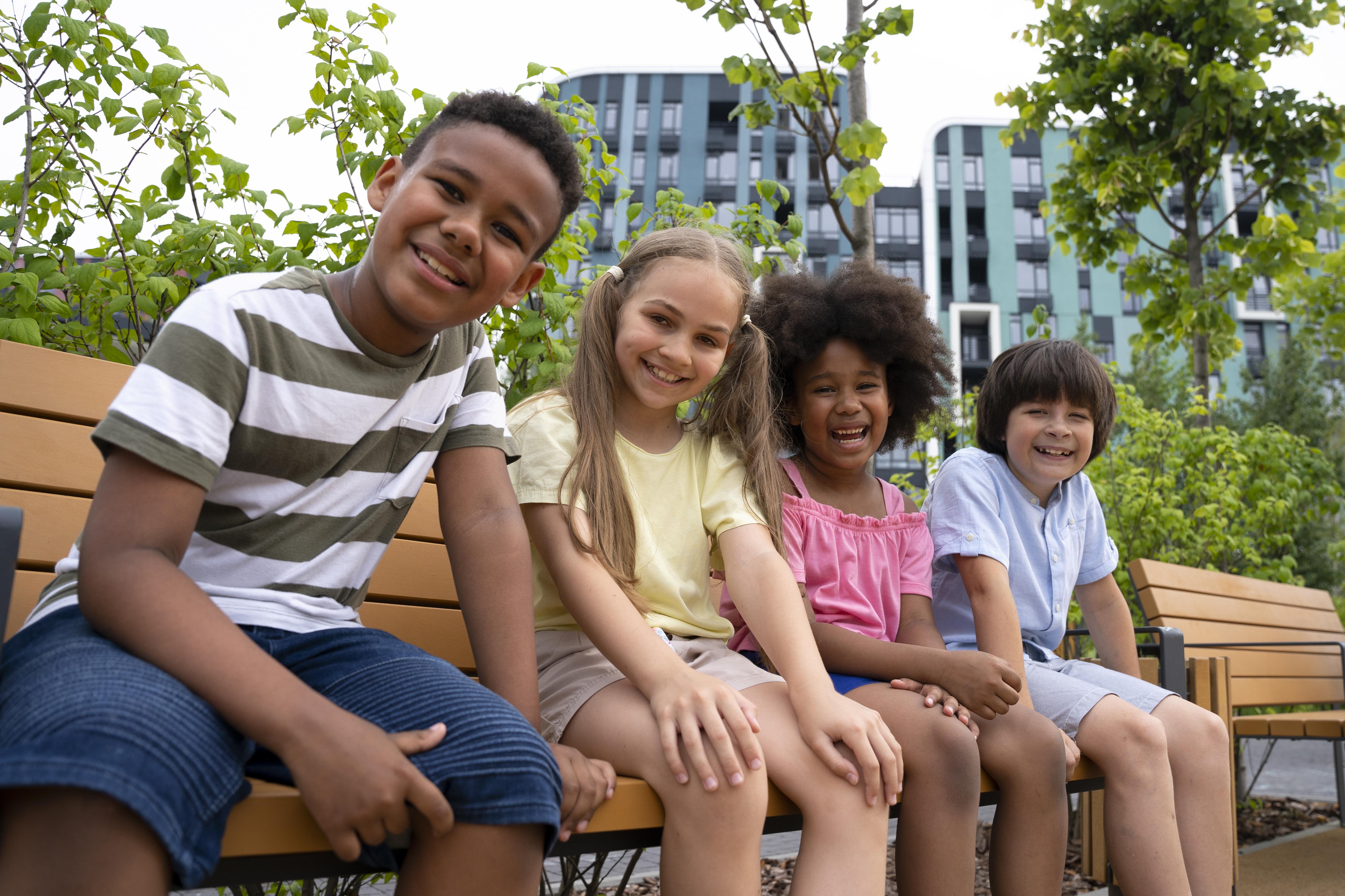 Four children sitting on a bench in an urban park with buildings in the background